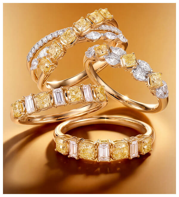 Four yellow gold rings with fancy yellow and white diamonds, shown stacked and overlapping on a warm gold background.