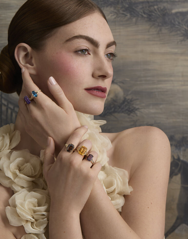 Model in a cream floral dress wearing bold cocktail rings with blue, purple, yellow and brown gemstones in vintage-inspired gold settings.