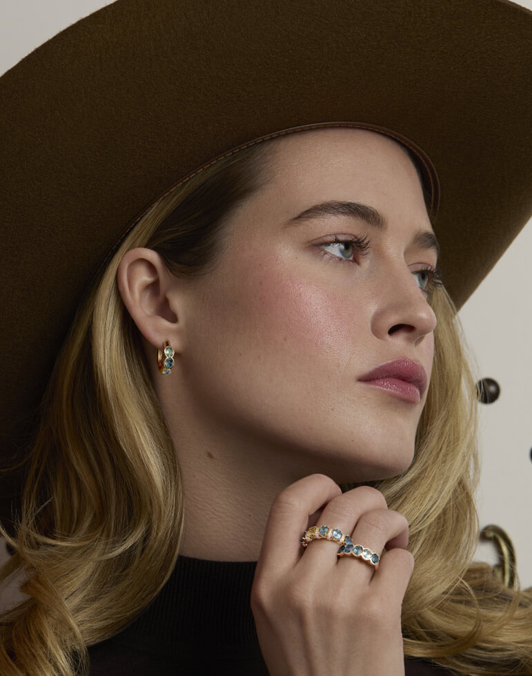 Blonde model in a wide-brim hat wearing blue-green gemstone earrings and rings in yellow gold, styled with a black top.
