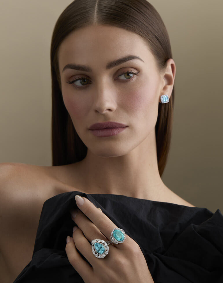 Model in a black off-the-shoulder top wearing large turquoise-colored gemstone rings and matching stud earrings with white diamond halos.